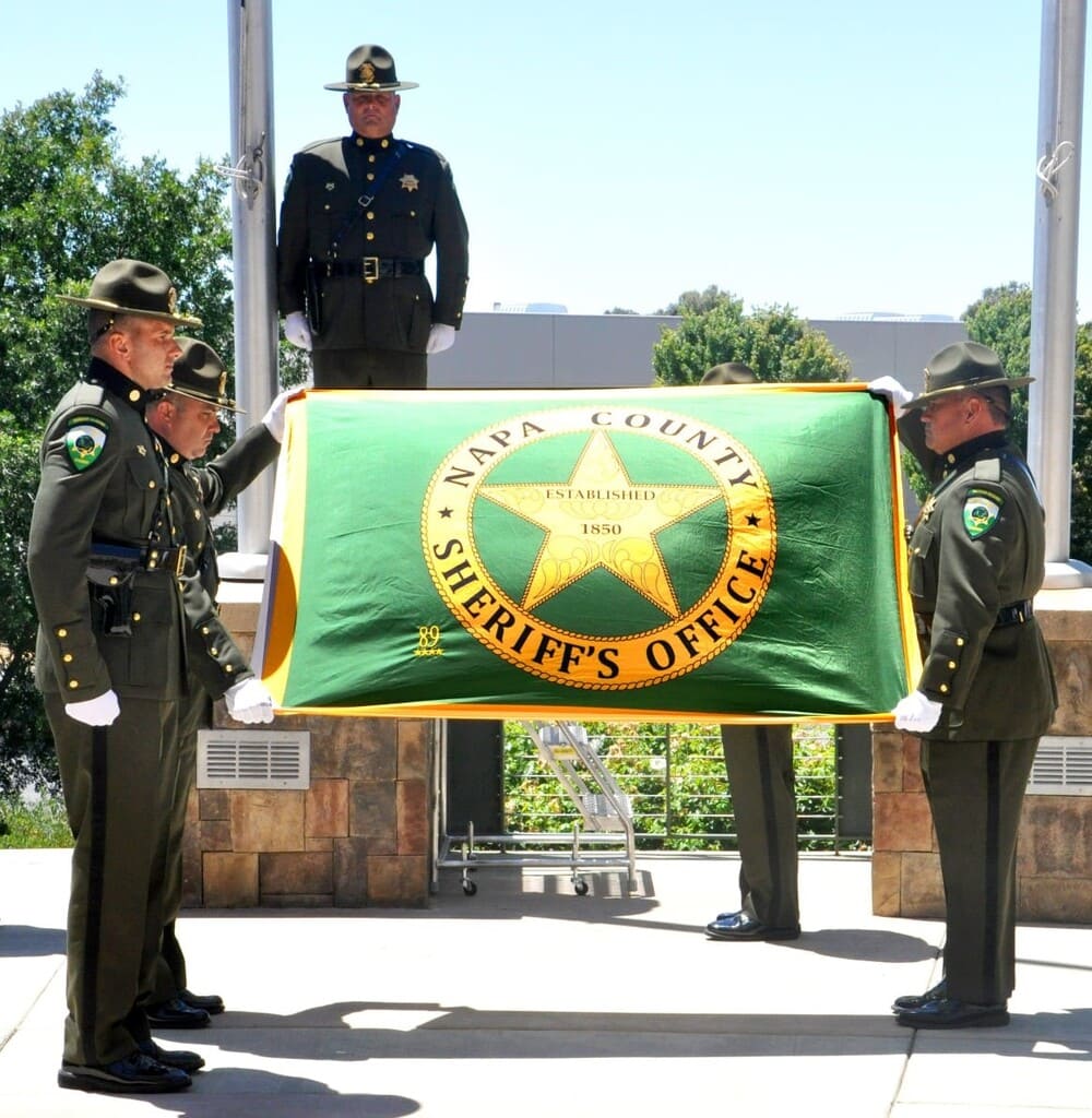 Four uniformed sheriff deputies holding a Napa County Sheriff's Office flag outdoors during a ceremony.