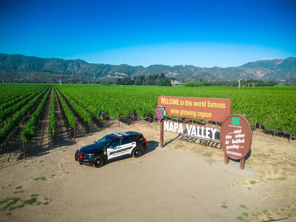 A parked Napa County sheriff SUV near a wooden Napa Valley sign welcoming visitors to the wine-growing region, surrounded by green vineyard rows under a clear blue sky.