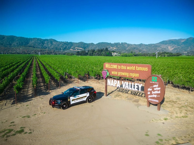 Napa Valley welcome sign beside vineyard rows with a sheriff's SUV parked on dirt under clear blue sky.