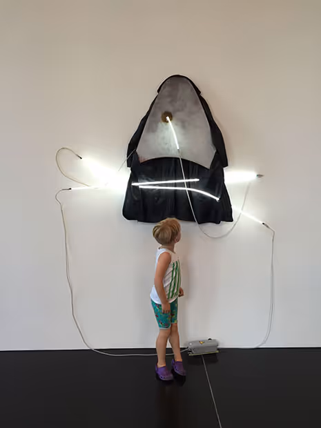 Young girl infront of an old coat hung on a wall with white neon lines cutting through it.