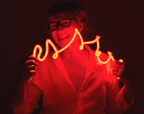A bio pic of Sue Shepherd. She is holding a string of neon that is casting a red glow across her face
