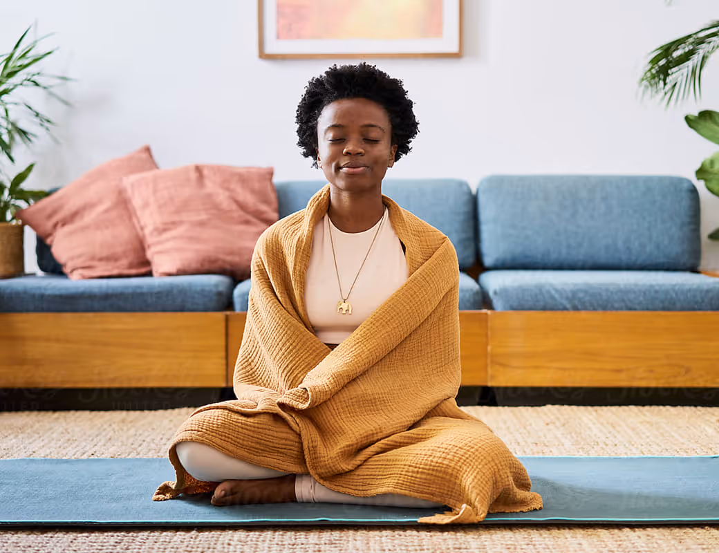 Woman meditating at home wrapped in a blanket on a yoga mat.