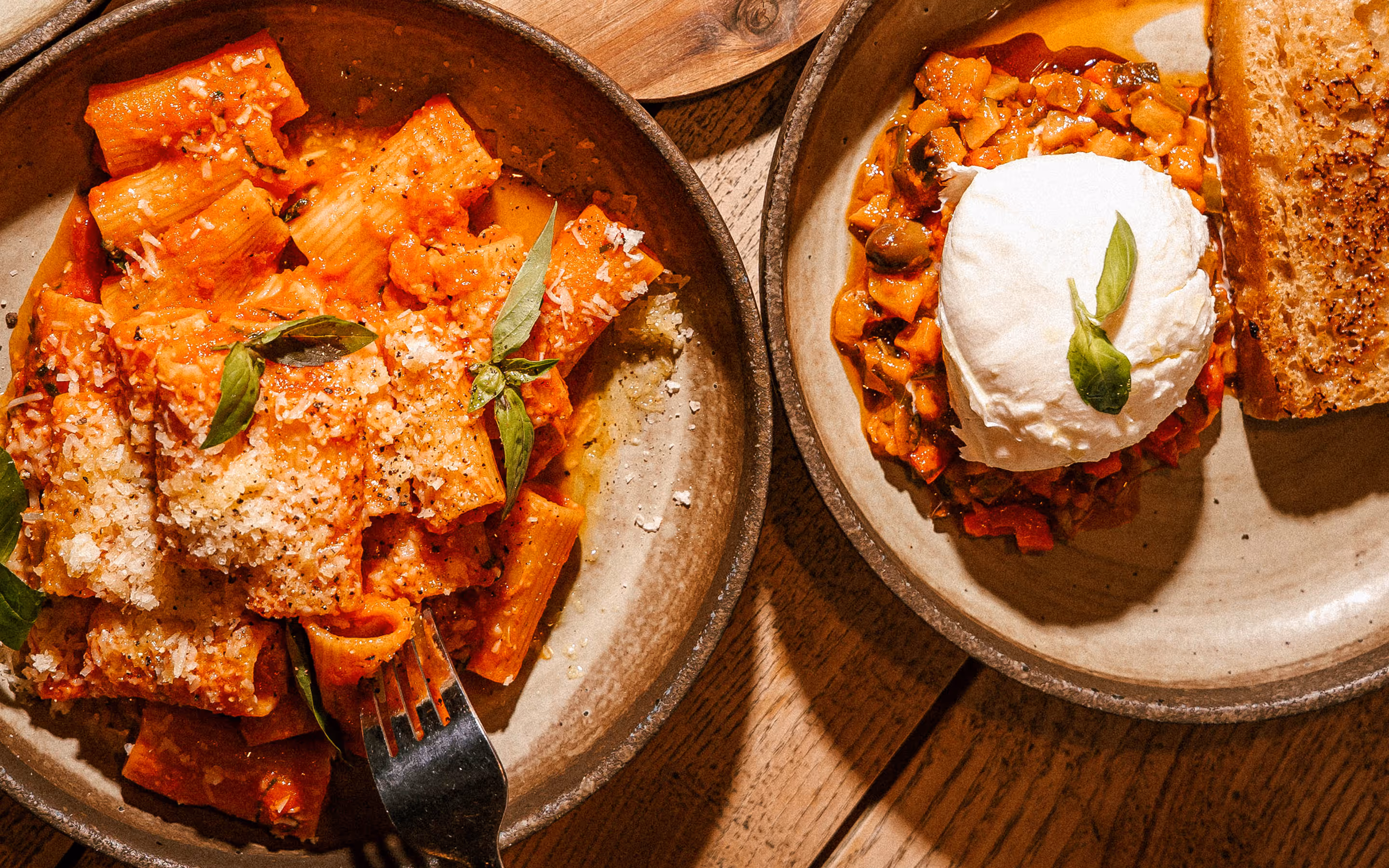 Talíř rigatoni těstovin s rajčatovou omáčkou, bazalkou a strouhaným sýrem vedle misky s mozzarellou, zeleninovou směsí a plátkem opečeného chleba na dřevěném stole.