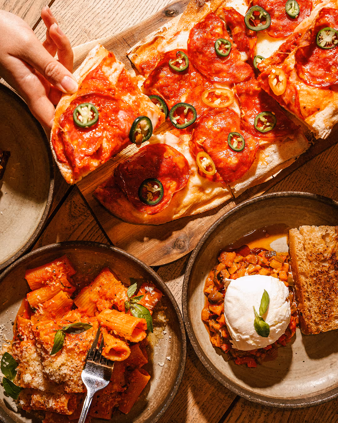 Ruka bere plátek pikantní pizzy s salámem a jalapeños, vedle talíř s těstovinami rigatoni a talíř s pečenou zeleninou, mozzarellou a kouskem opečeného chleba.