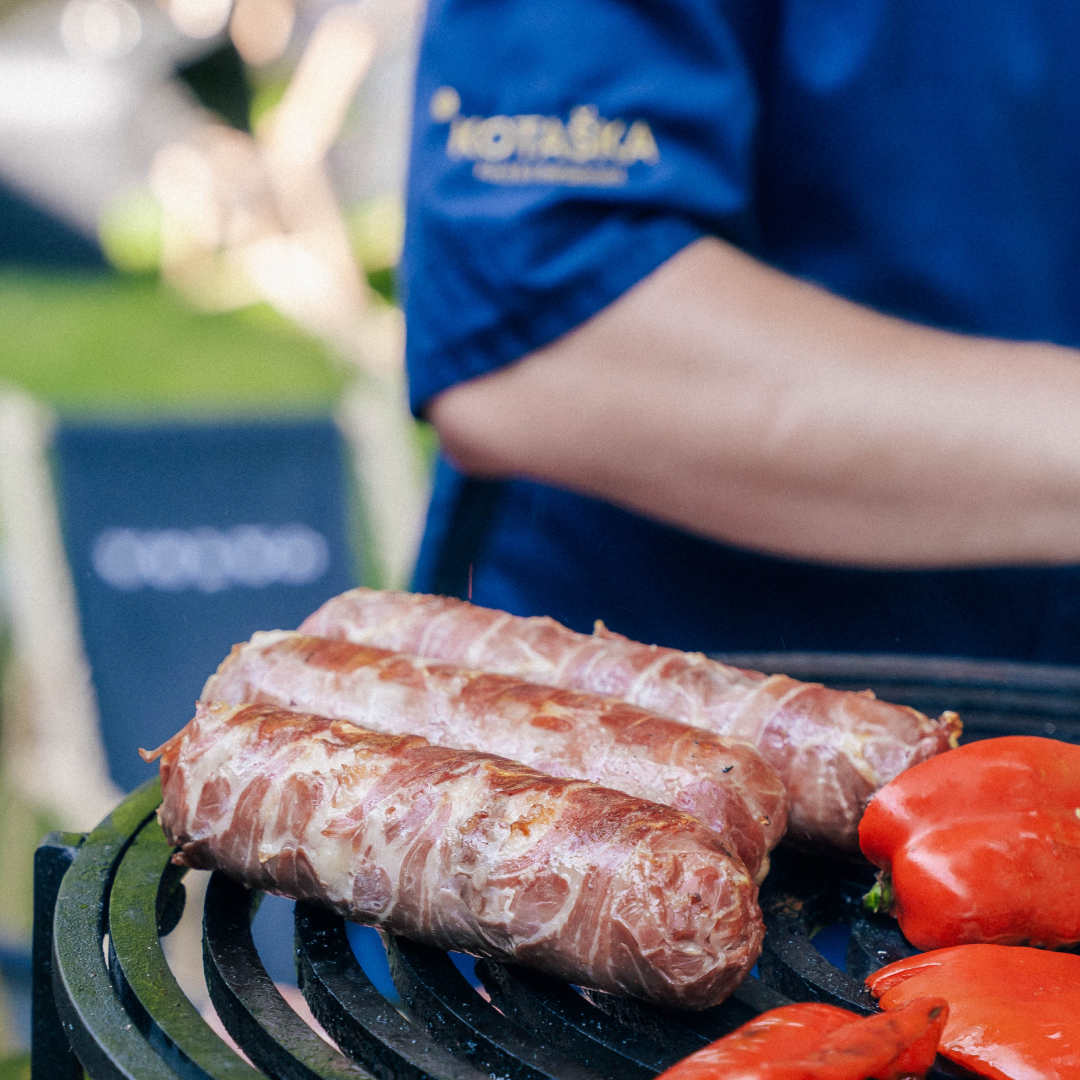 Grilované klobásy a červené papriky na černém grilu s rukou v modrém tričku v pozadí.
