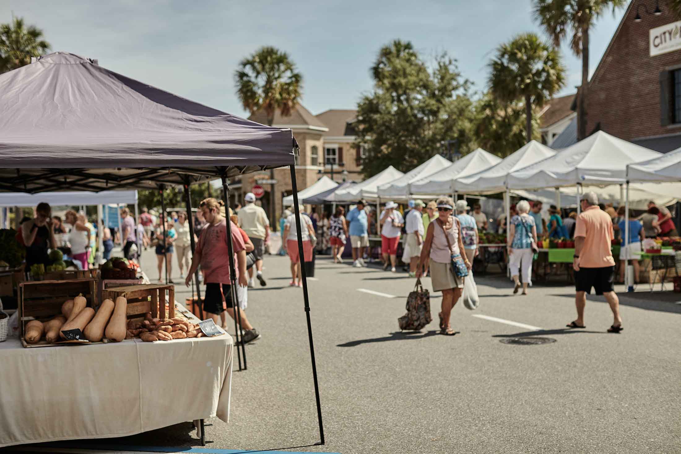 People walking and shopping at an outdoor farmers market with tents and fresh produce on display under sunny weather.