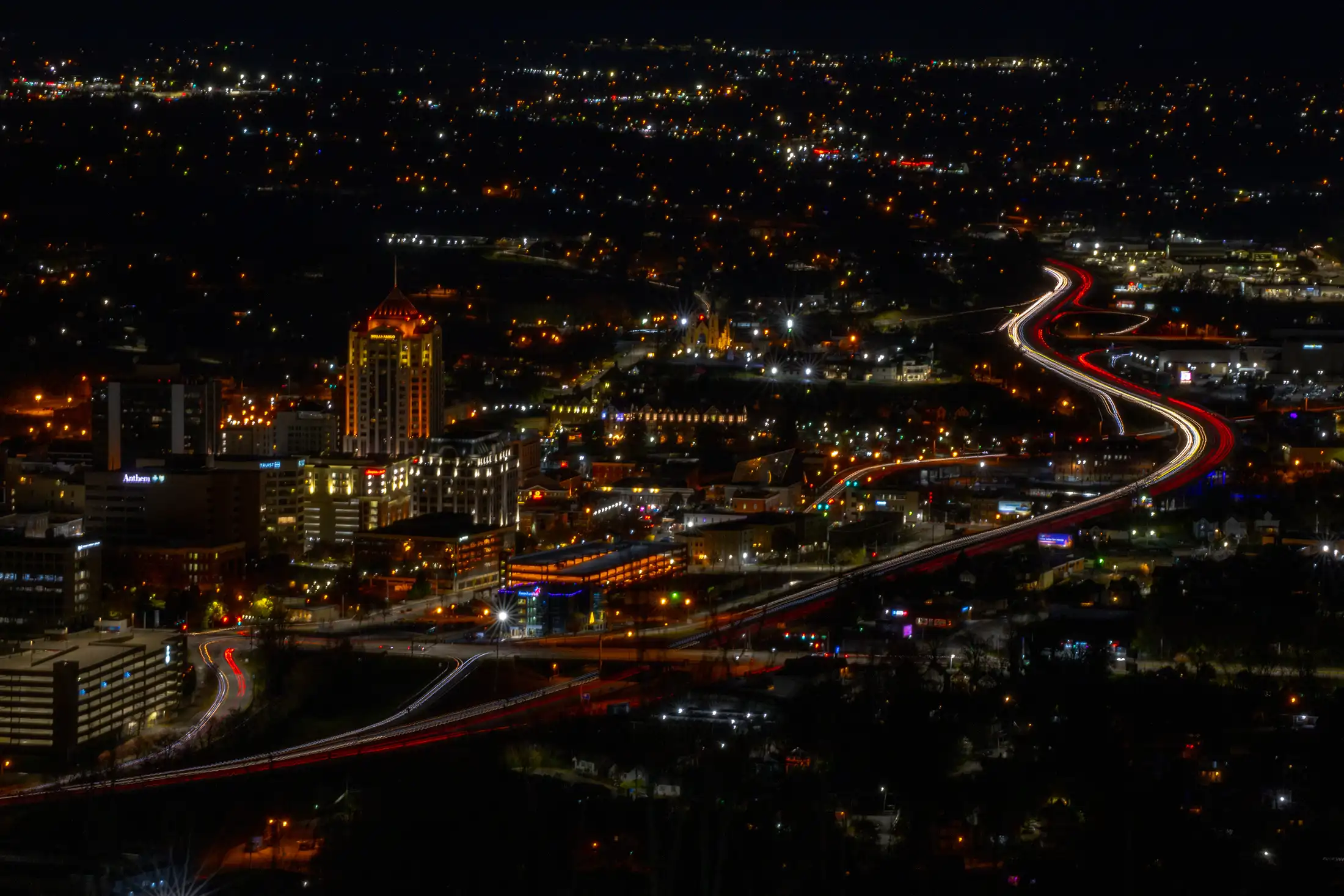 Aerial night view of a city skyline with illuminated buildings and light trails from cars on winding highways.