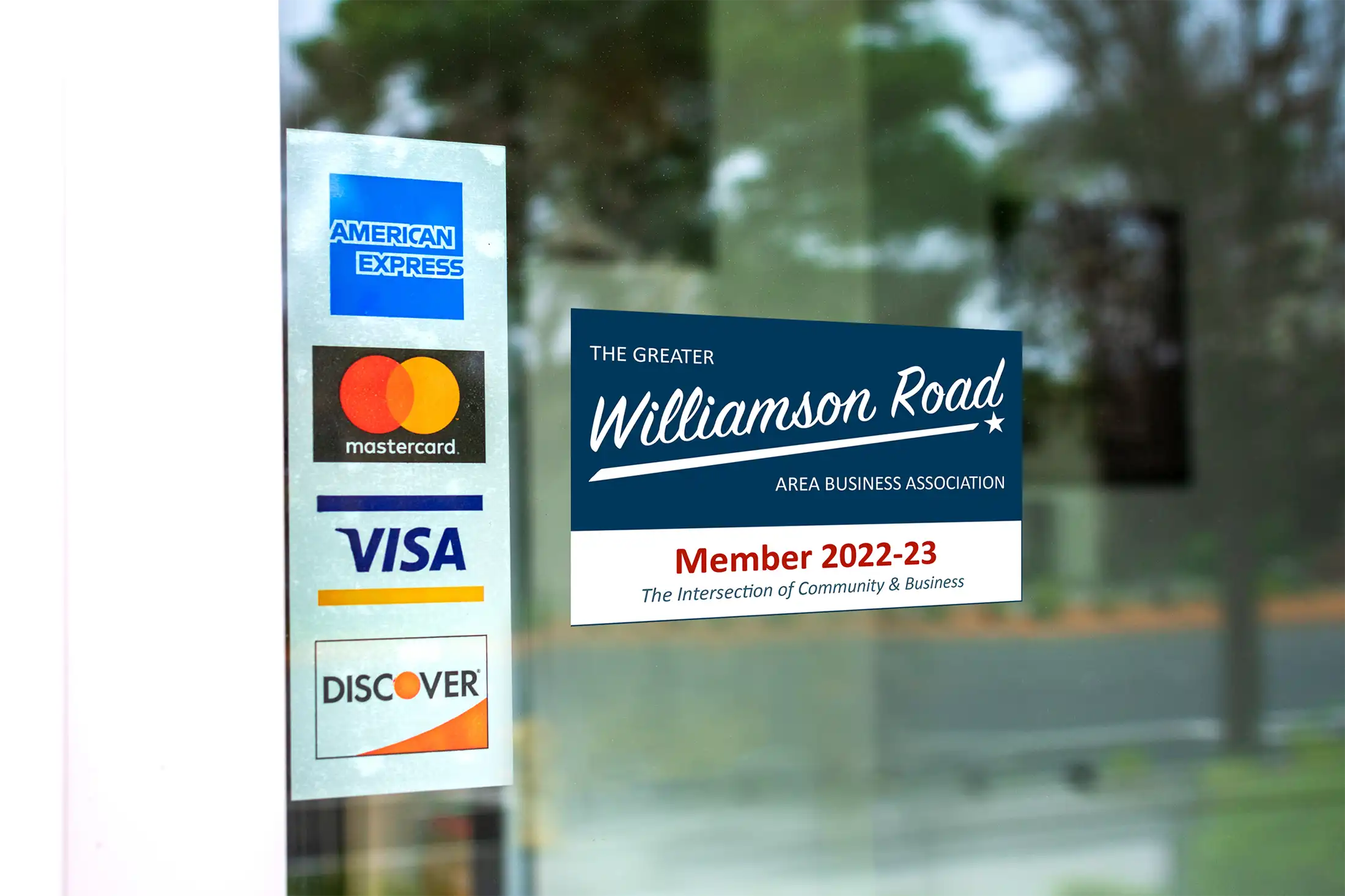 Window stickers showing accepted payment cards American Express, Mastercard, Visa, Discover, and a membership decal for The Greater Williamson Road Area Business Association for 2022-23.