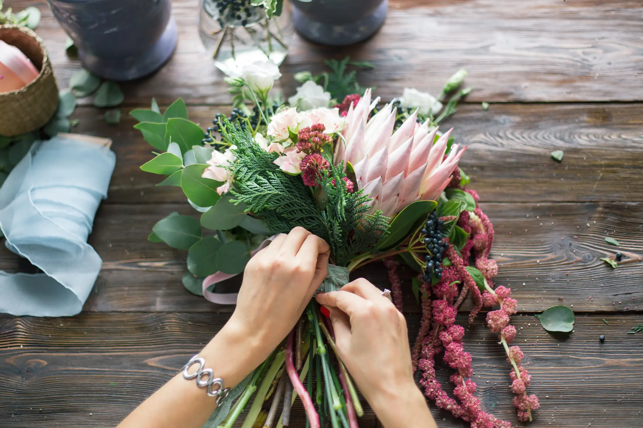 Hands arranging a bouquet with pink protea, greenery, and other flowers on a wooden surface.
