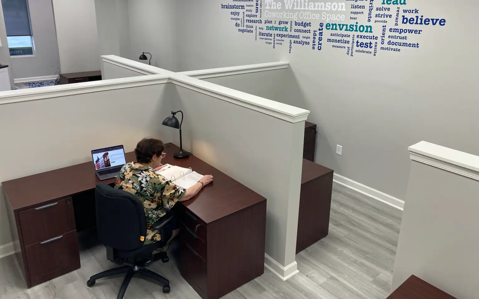 Person sitting at a desk in a cubicle, reading a binder with a laptop and desk lamp nearby, in a modern office space.