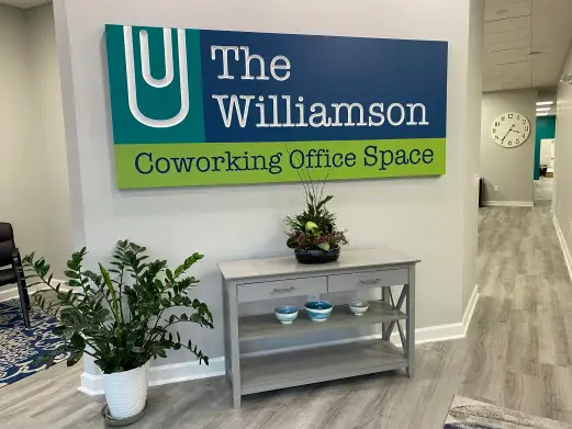 Entry area of The Williamson coworking office space with a sign on the wall, a gray console table with decorative bowls and plants, and light wood flooring.