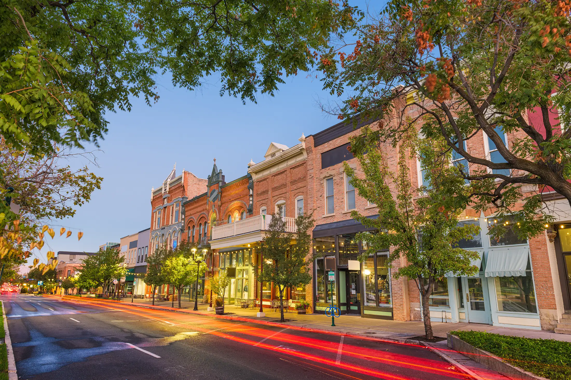 Street view of historic brick buildings with storefronts and trees at dusk, with red light trails from passing cars.