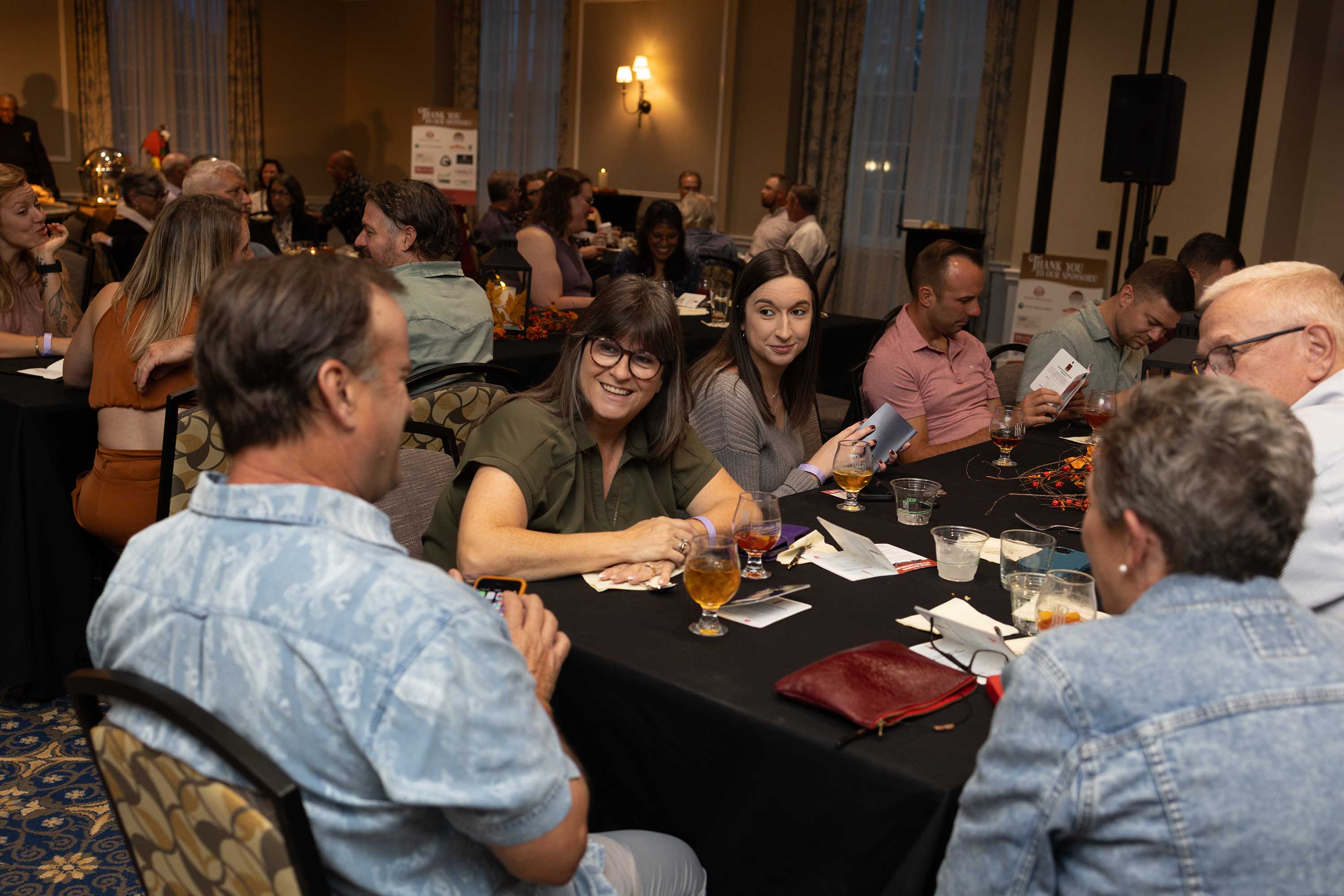 Group of people seated around a table at an indoor social event, engaged in conversation and holding drinks.