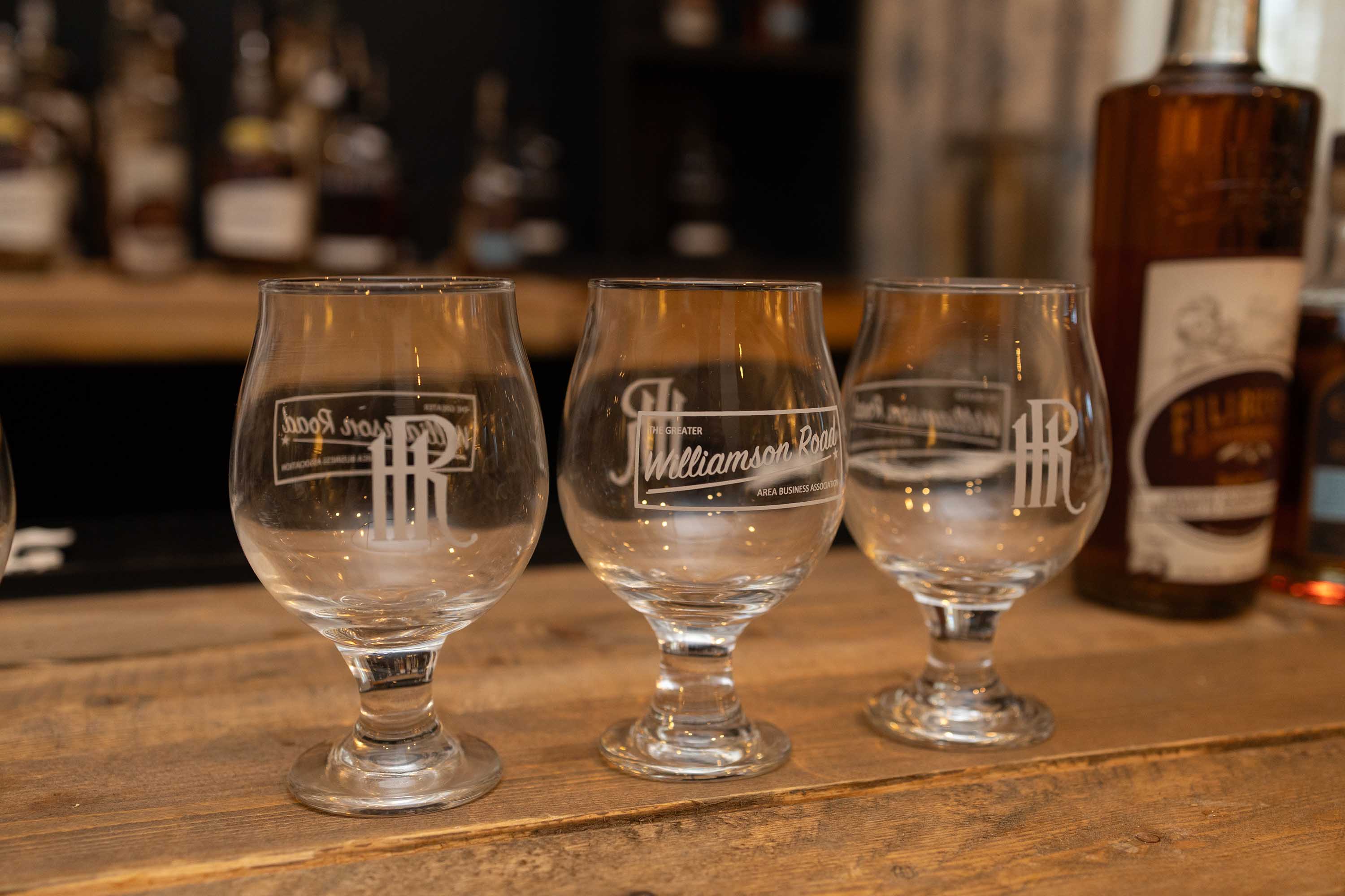 Three empty tulip-shaped whiskey glasses with 'The Greater Williamson Road Area Business Association' logo on a wooden surface, with bottles blurred in the background.