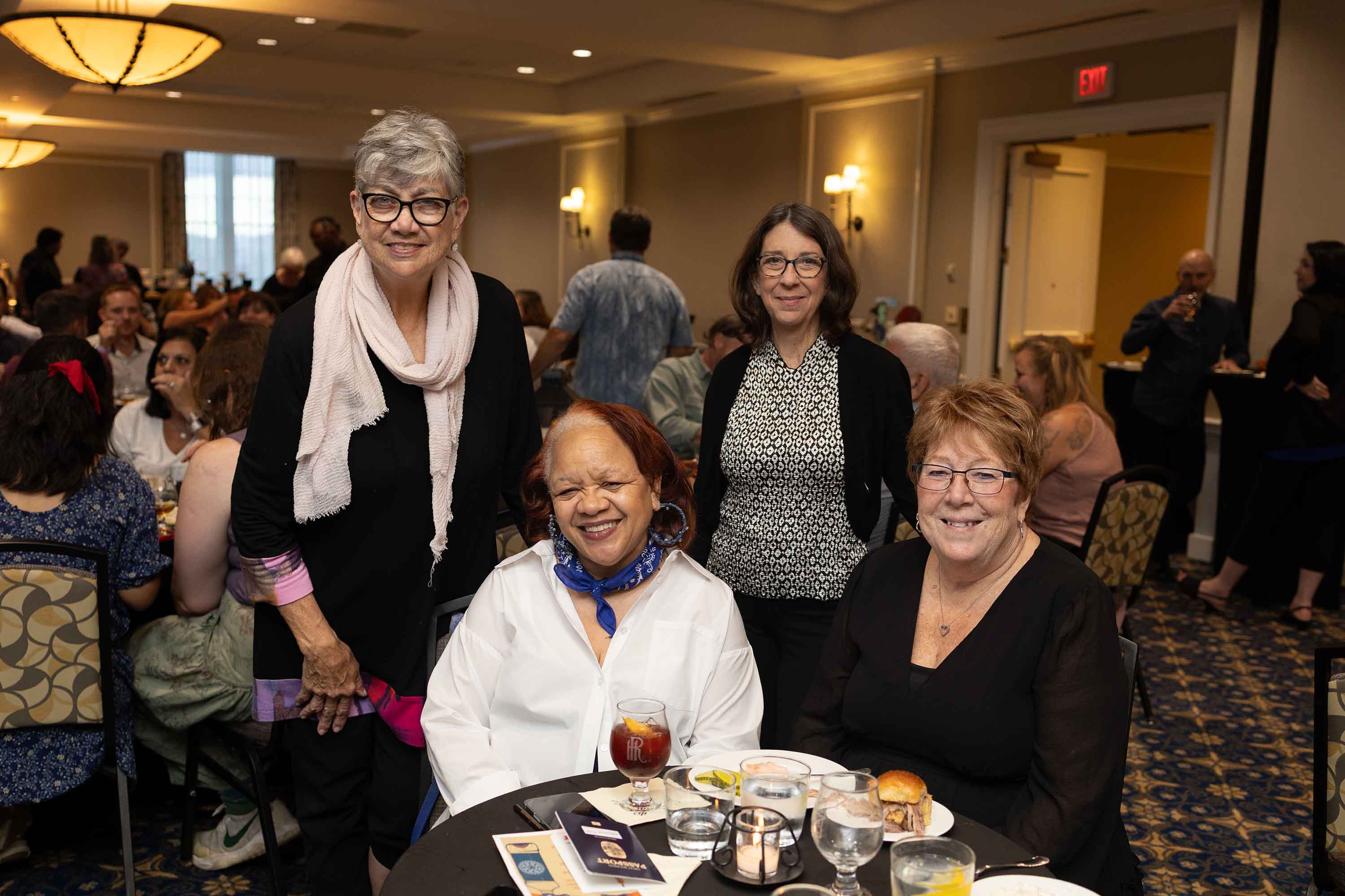 Four women smiling at a social gathering, with two seated and two standing behind them at a table set with drinks and food.