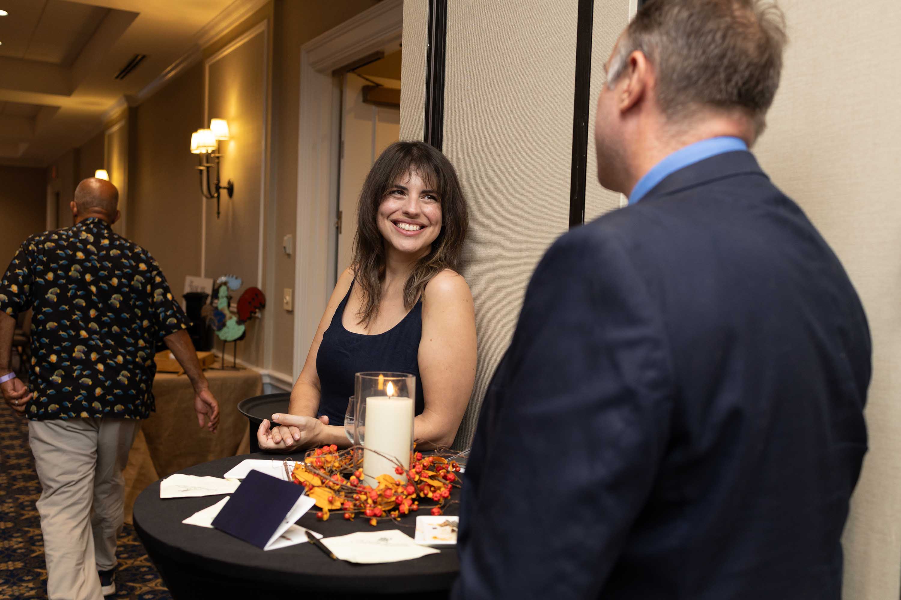 Smiling woman in a black dress talking to a man in a navy suit near a candle-decorated table in a hallway.