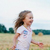 A young girl with blonde hair and a pink shirt is smiling and running in a field.