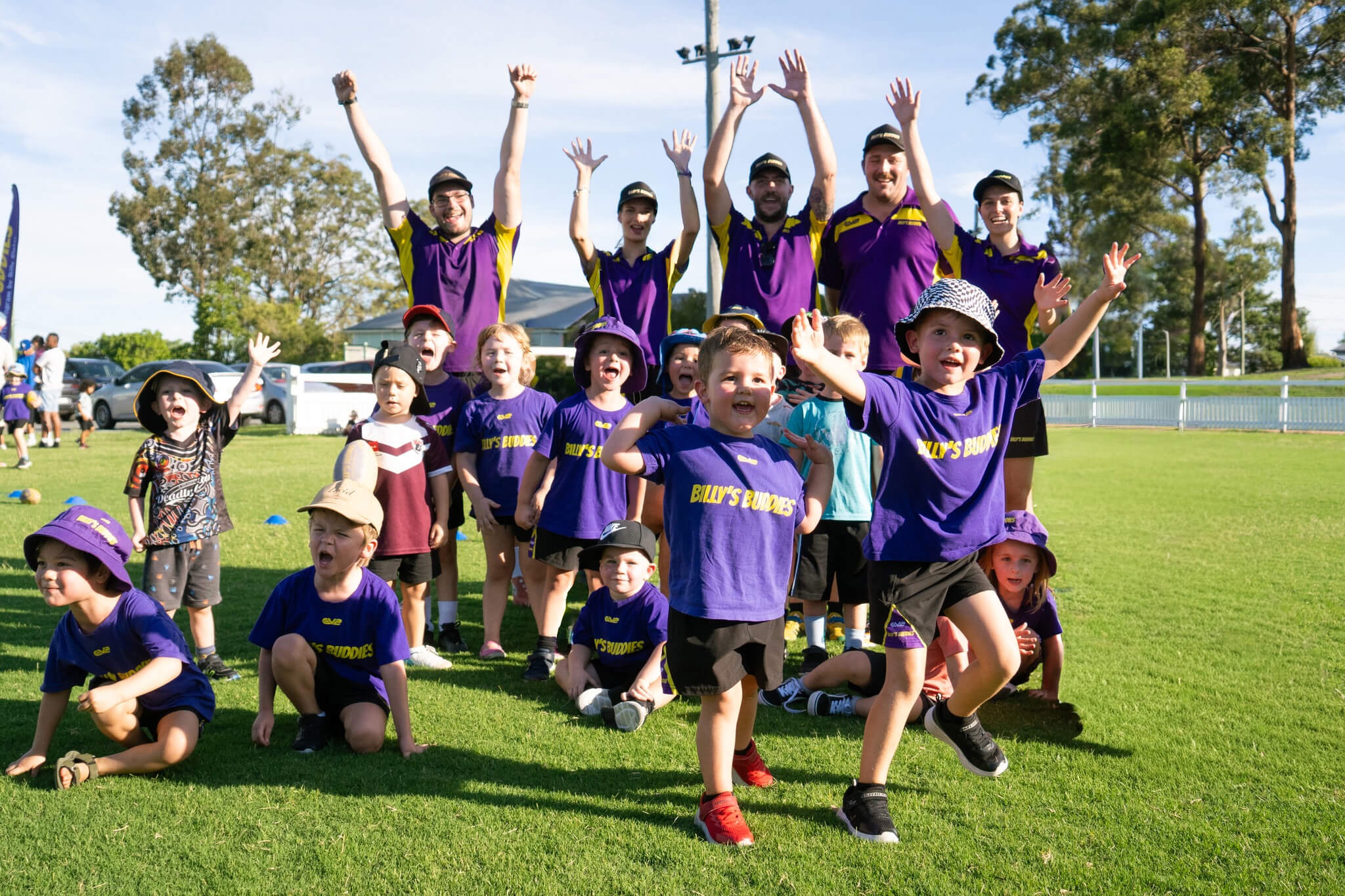 Group of children in purple 'Billy's Buddies' shirts and adults in matching polo shirts raising their hands outdoors on a sunny sports field.