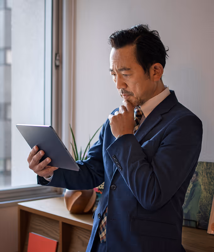 Mature businessman in a navy suit thoughtfully reviewing content on a tablet near a window in a modern office. Ideal for concepts like digital leadership, executive decision-making, or professional remote work.