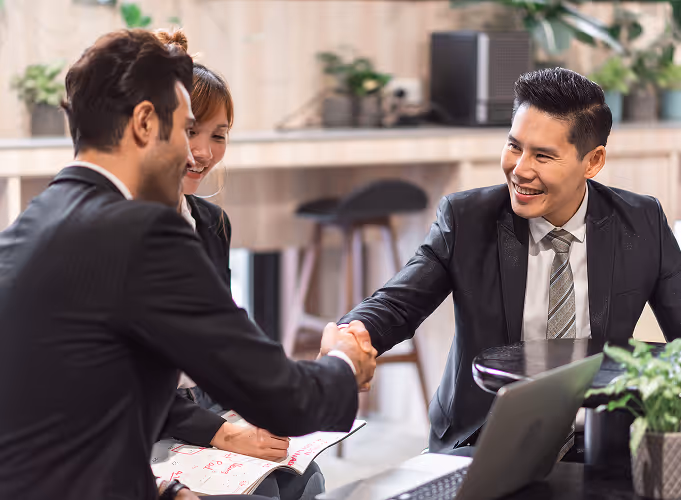 Business professionals in formal attire shaking hands across a desk in a modern office, symbolizing partnership, successful negotiation, or client onboarding. Smiling team members and open laptops reflect a productive and collaborative business meeting.