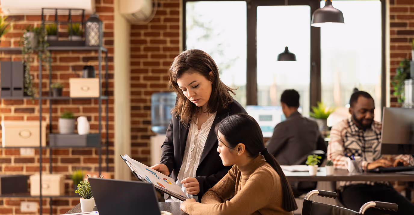 Female team leader mentoring a colleague in a modern coworking office with exposed brick walls and natural light. Business charts and laptops highlight productivity, teamwork, and professional development in a collaborative workplace setting.