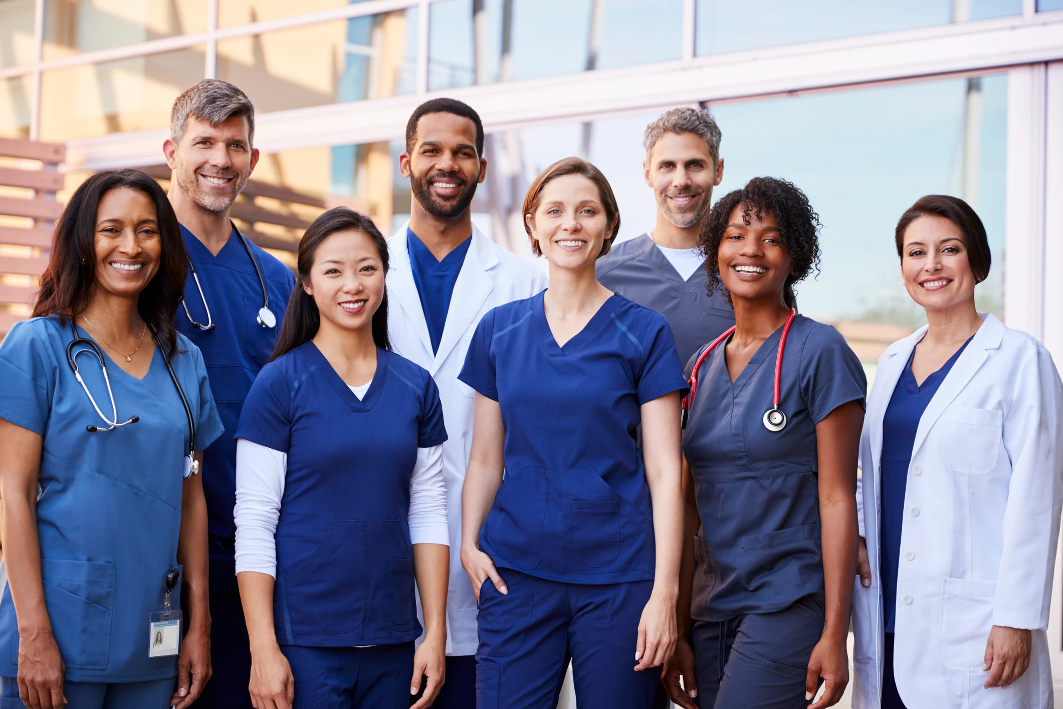 Group of smiling diverse healthcare professionals wearing scrubs and lab coats posing outside a modern building.