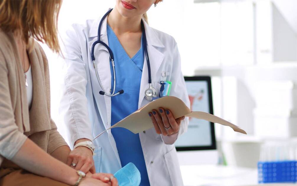 Female doctor in white coat and blue scrubs holding a file and talking to a seated female patient.