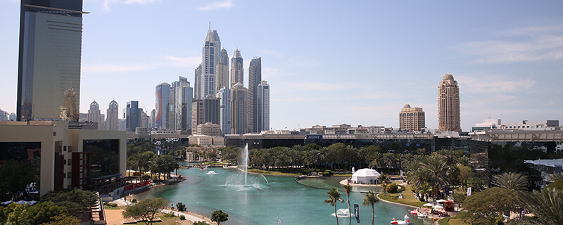 Cityscape with tall modern skyscrapers behind a large water fountain in a landscaped park.
