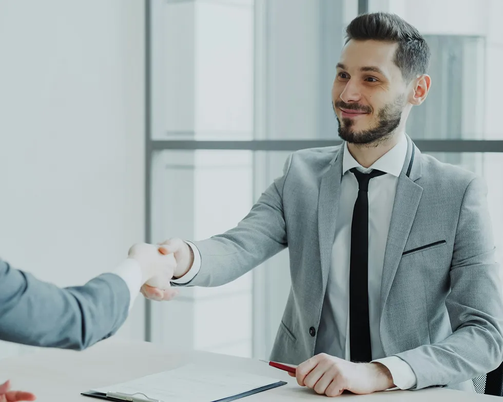 Businessman in a gray suit shaking hands with another person across a desk with documents.