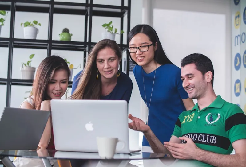 Four diverse young professionals gathered around a laptop, collaborating and discussing work in a modern office setting.