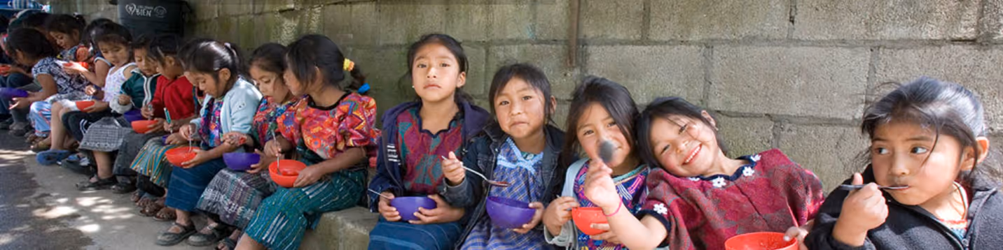 More than a dozen little Guatemalan girls sitting on a long bench outside, eating from bowls.