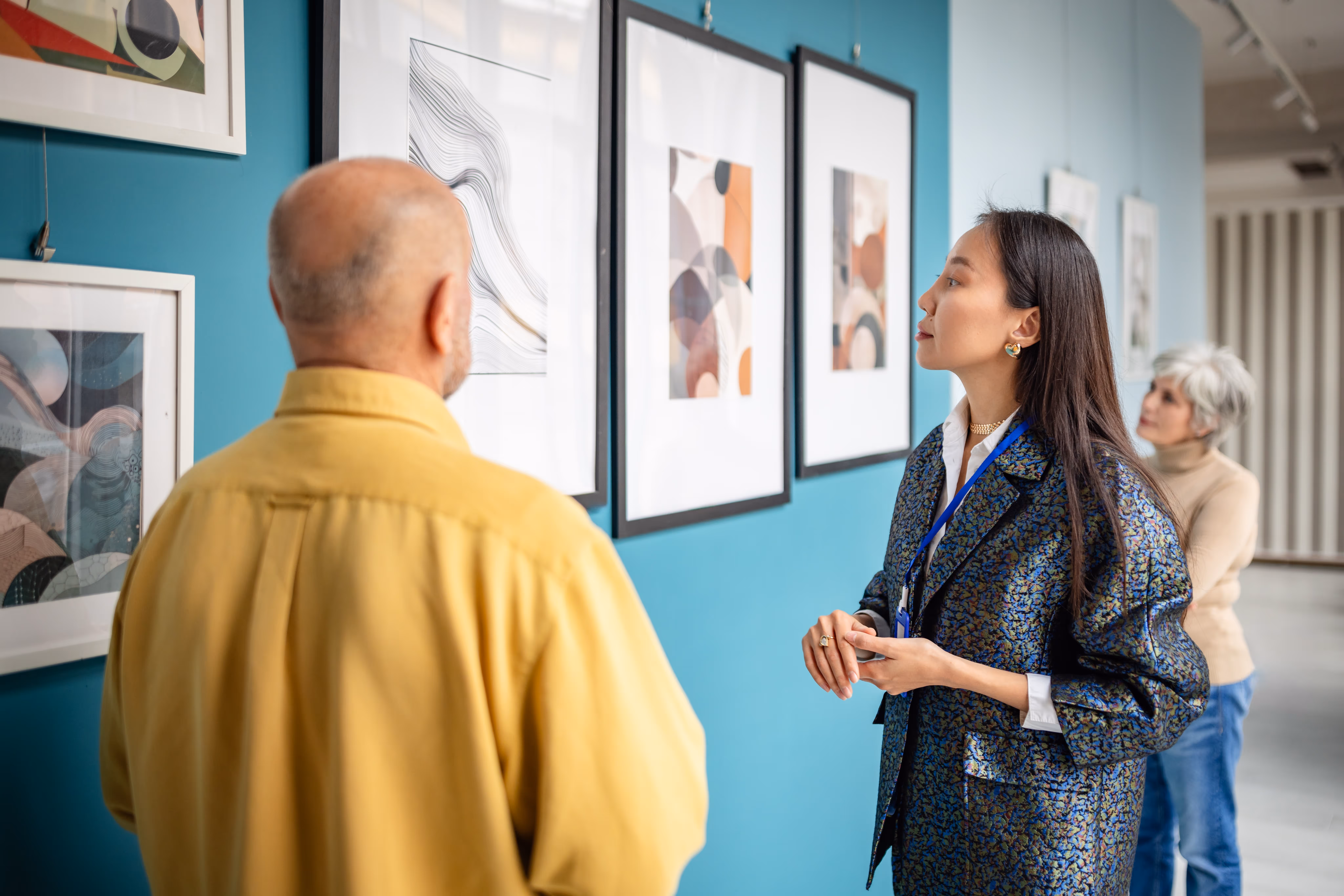 An older white couple talking to an Asian docent at an art gallery