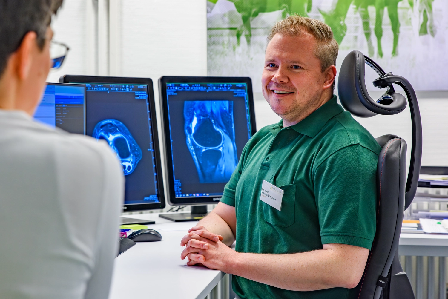 STENUM – Fachklinik für Orthopädie – Arzt berät einen Patienten an einem Schreibtisch mit zwei Monitoren, auf denen Röntgenbilder des Knies zu sehen sind.