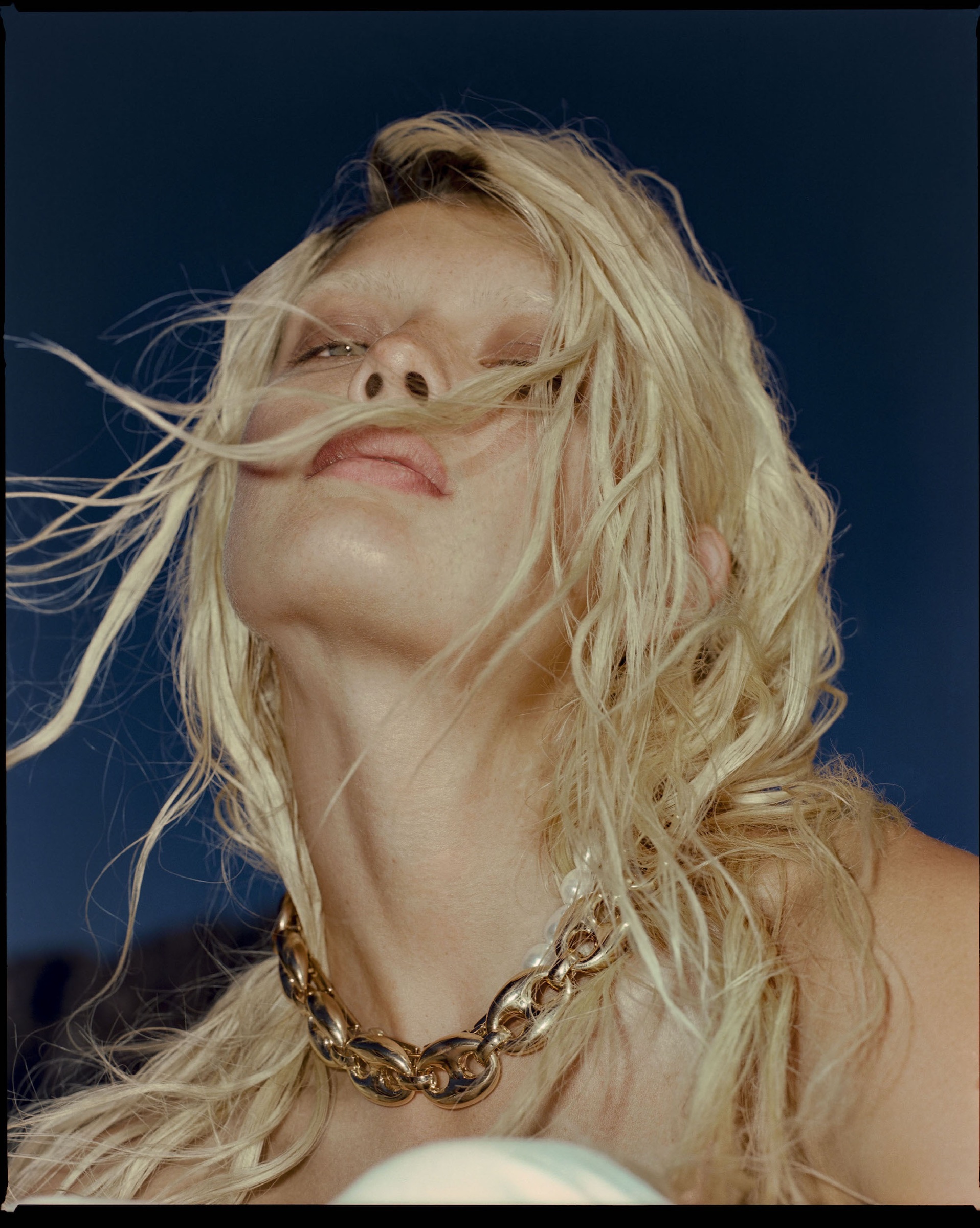 Model with windblown blonde hair and a heavy gold chain, head tilted back against a deep blue background