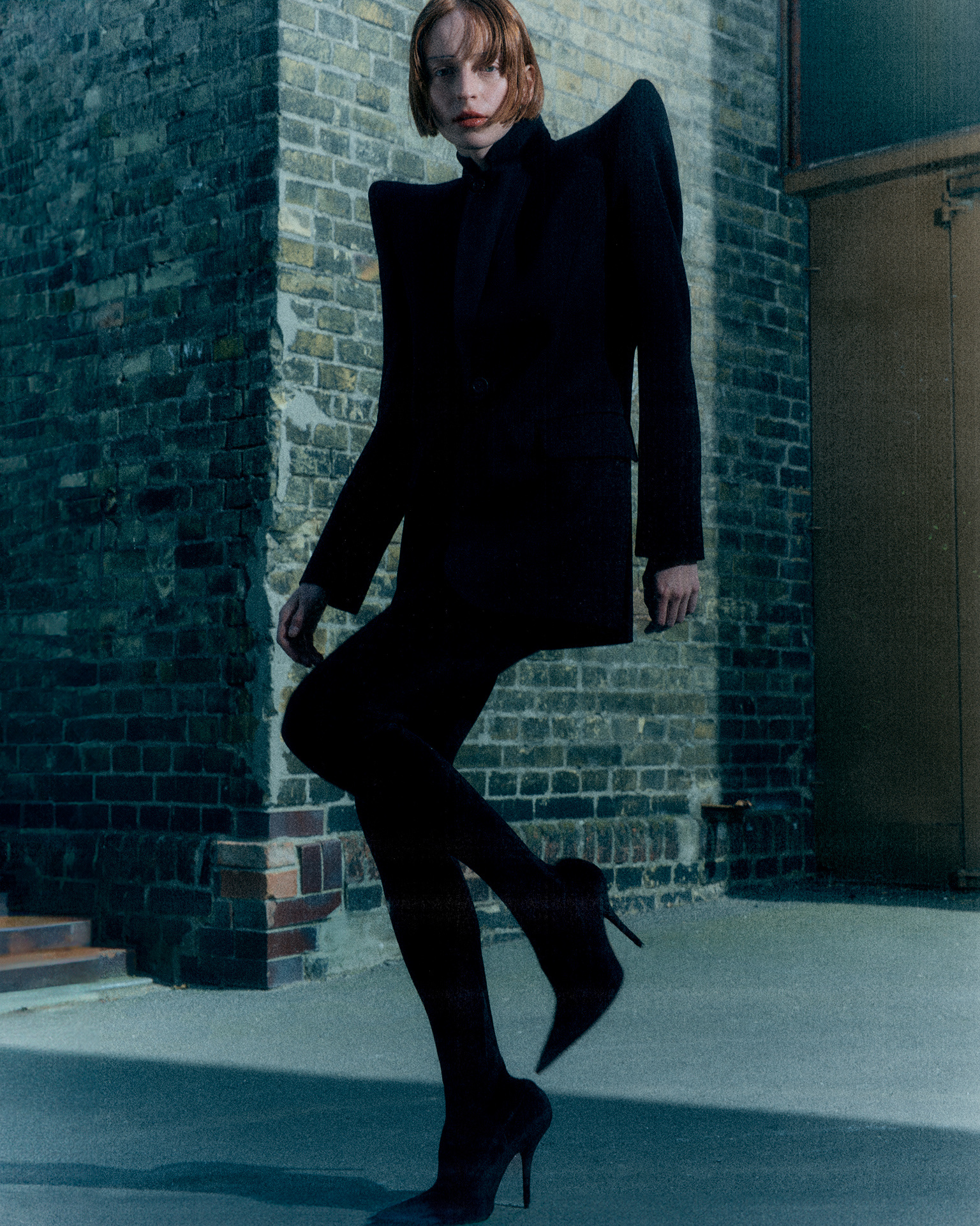 Model in a black jacket with wide padded shoulders posing against a brick wall in hard side light
