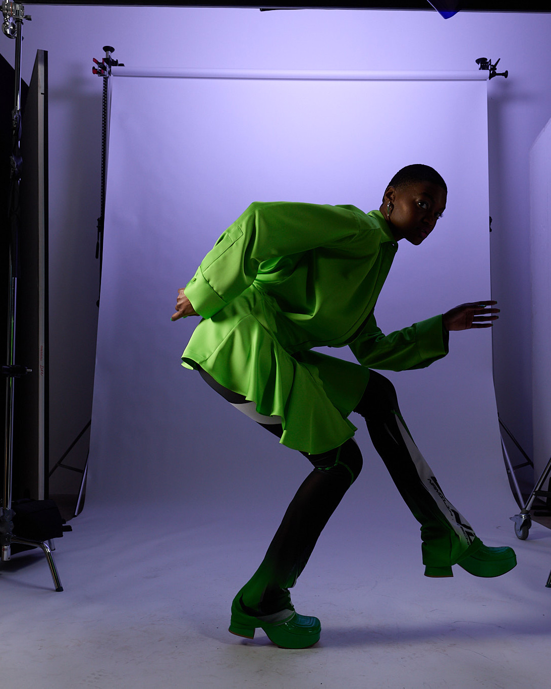 A model in a green jacket bending forward in front of a lilac backdrop, studio stands visible at the edges