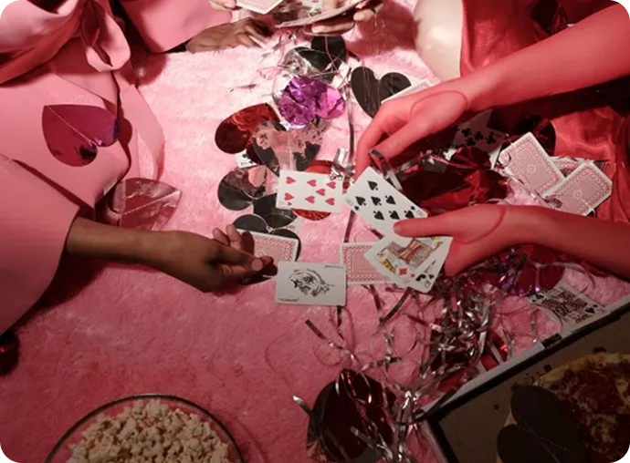 Two people in pink outfits playing cards on a pink surface surrounded by heart-shaped confetti, popcorn, and pizza.