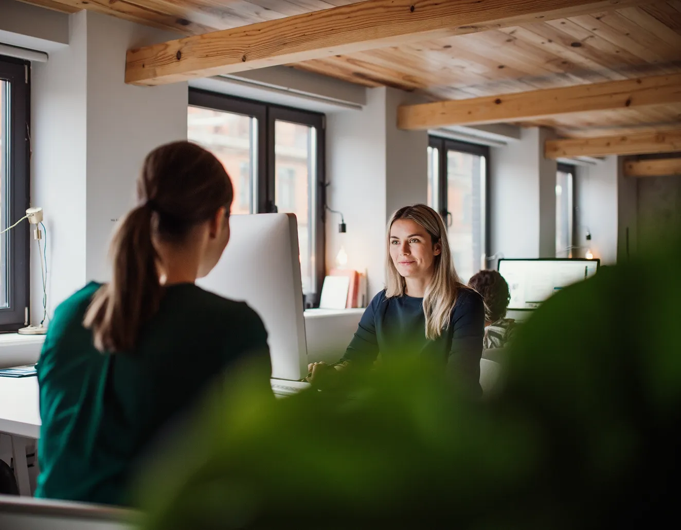 Two women talking at a desk in a modern office with wooden ceiling beams and large windows.