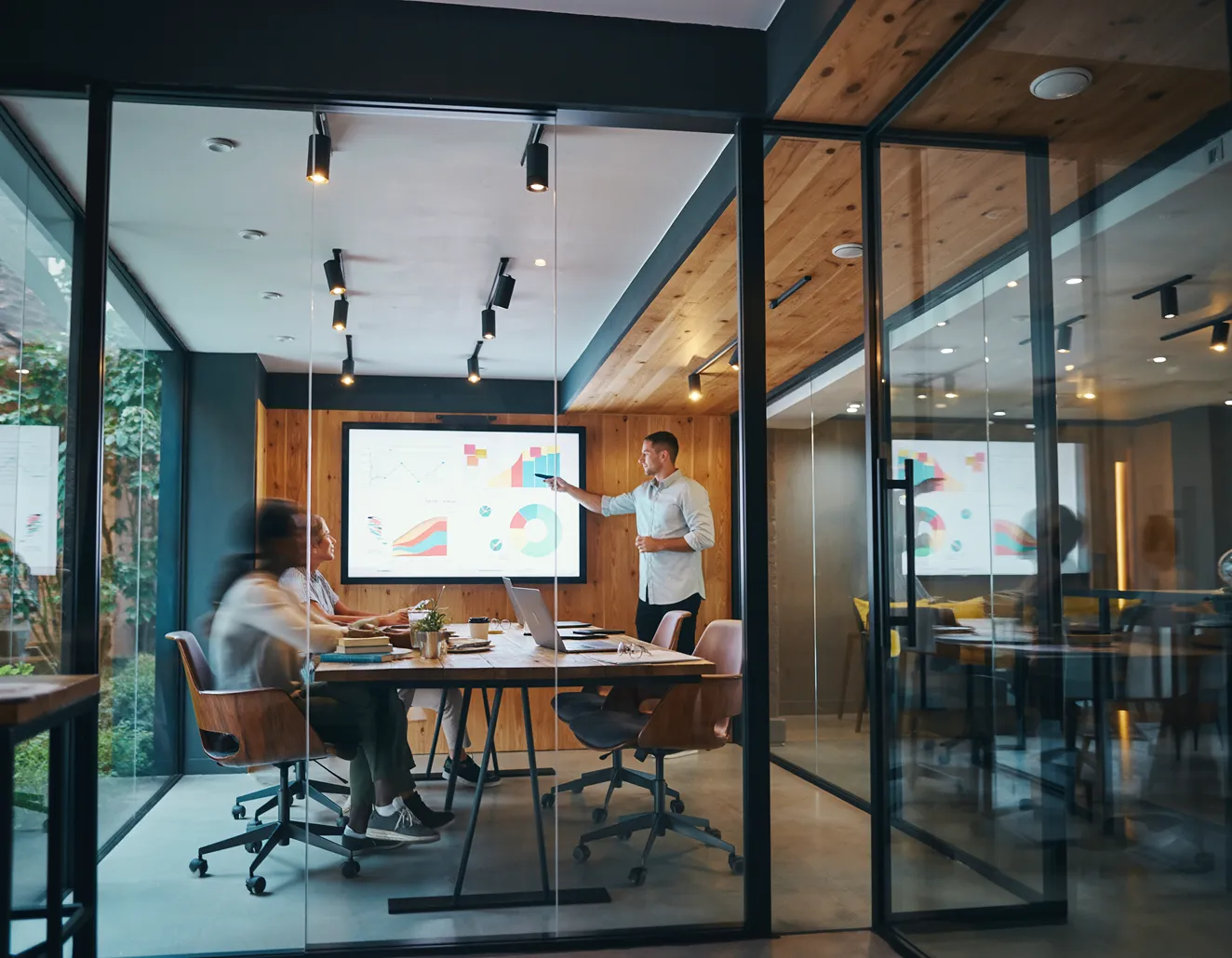 Man håller presentation med grafdiagram på en skärm för två personer i ett modernt konferensrum med glasväggar.