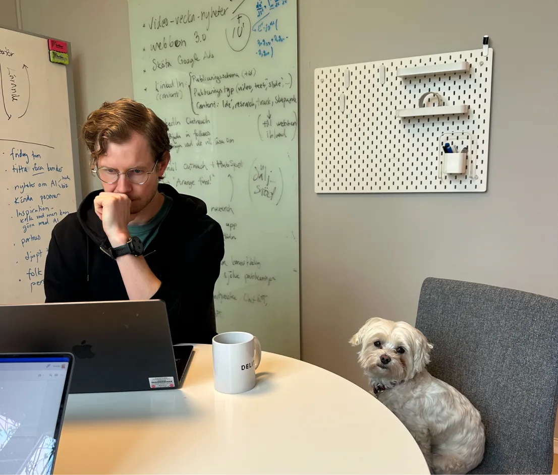 Man wearing glasses and a black hoodie working on a laptop at a table with a cup, and a small white dog sitting on a chair next to him.