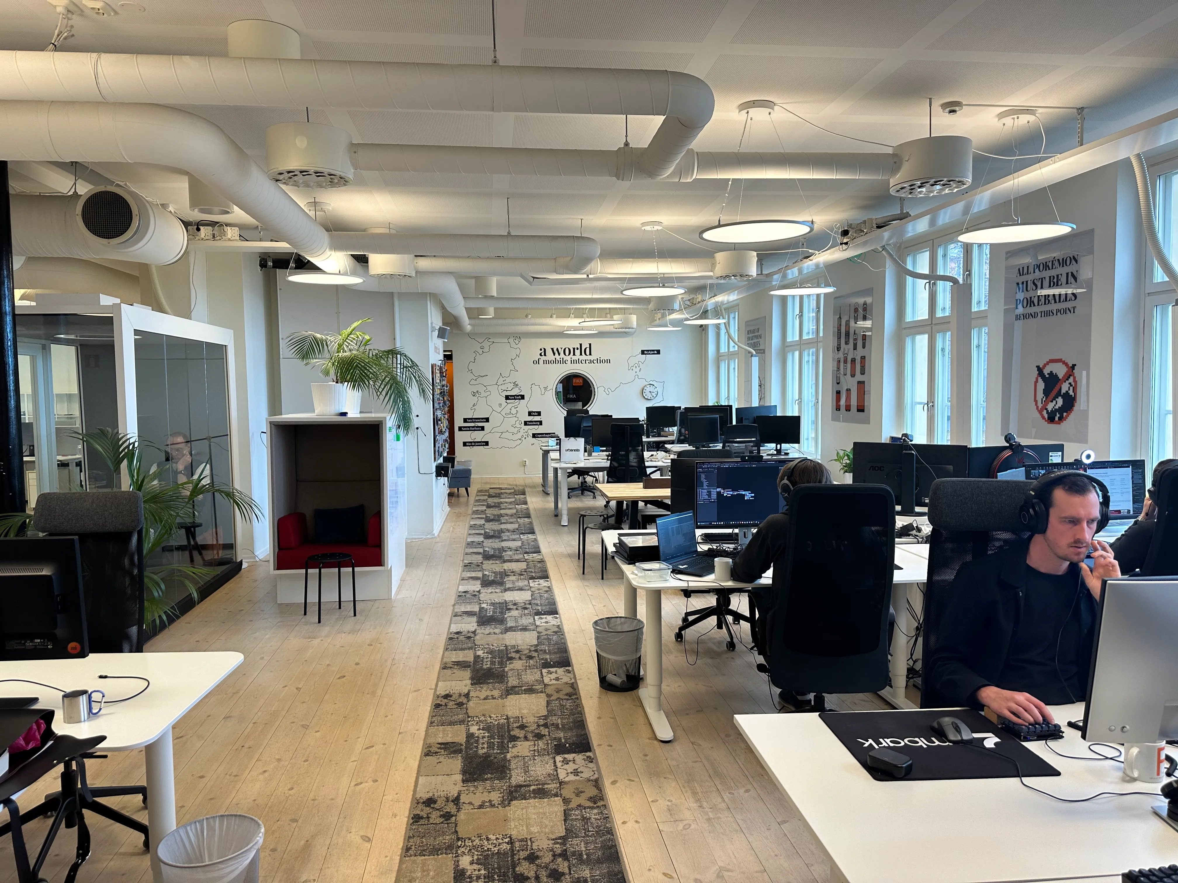 Modern open-plan office with wooden floors, multiple desks with computers, a carpeted walkway, and windows on the right side.