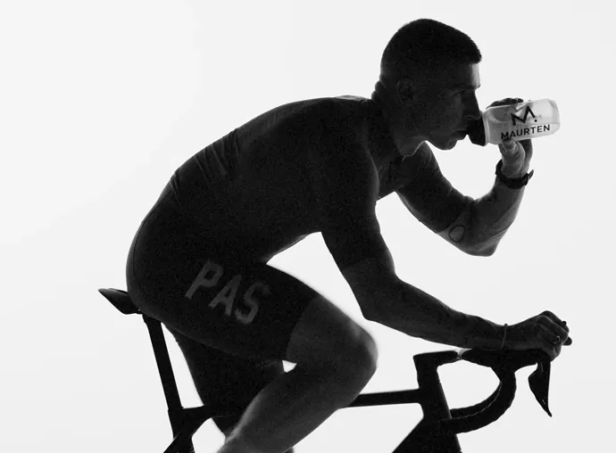 Silhouette of a cyclist drinking from a Maurten-branded bottle while riding a bike.