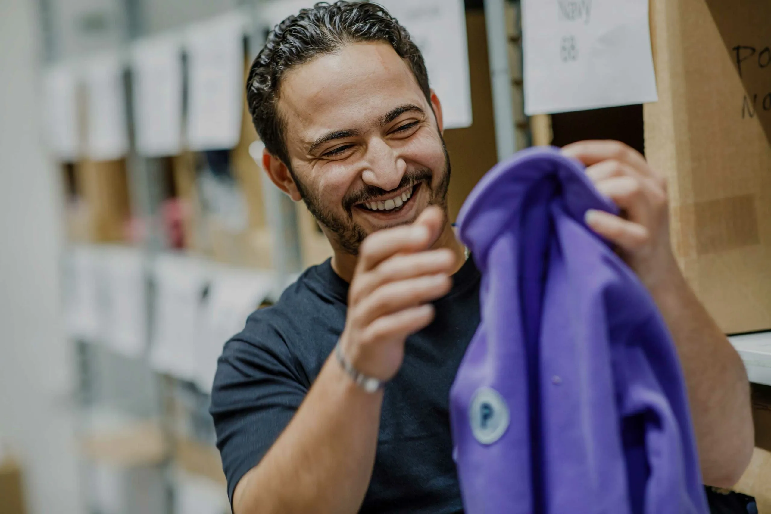 Smiling man holding and inspecting a purple jacket in a storage room with boxes on shelves.