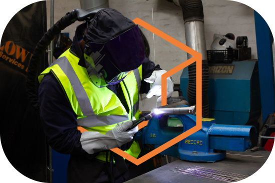 Worker wearing a welding helmet and high-visibility vest welding metal in a workshop.
