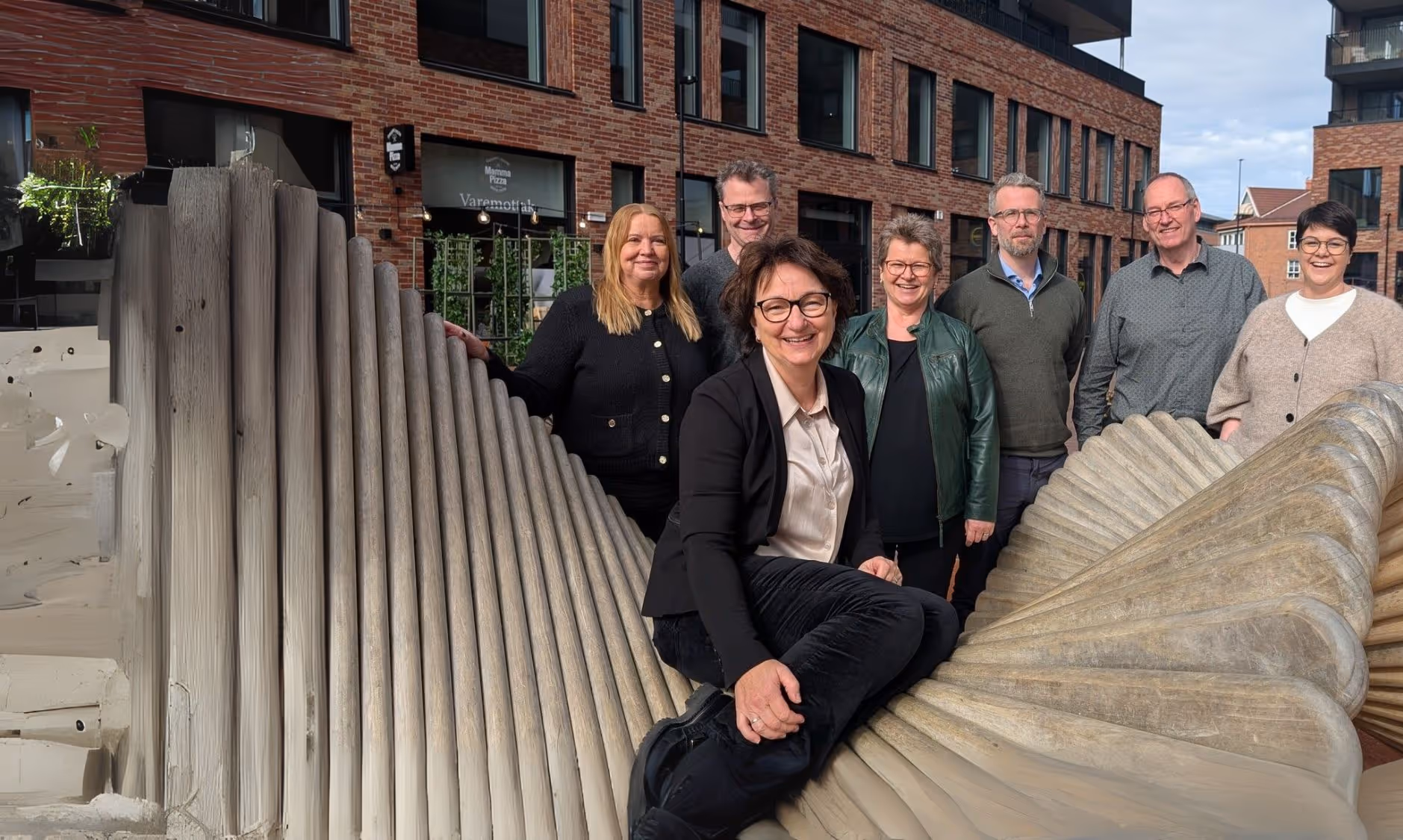 Åtte voksne poserer og smiler foran en moderne murbygning; en kvinne i forretningsklær sitter på en stor, riflet metallskulptur, mens de andre står bak henne.