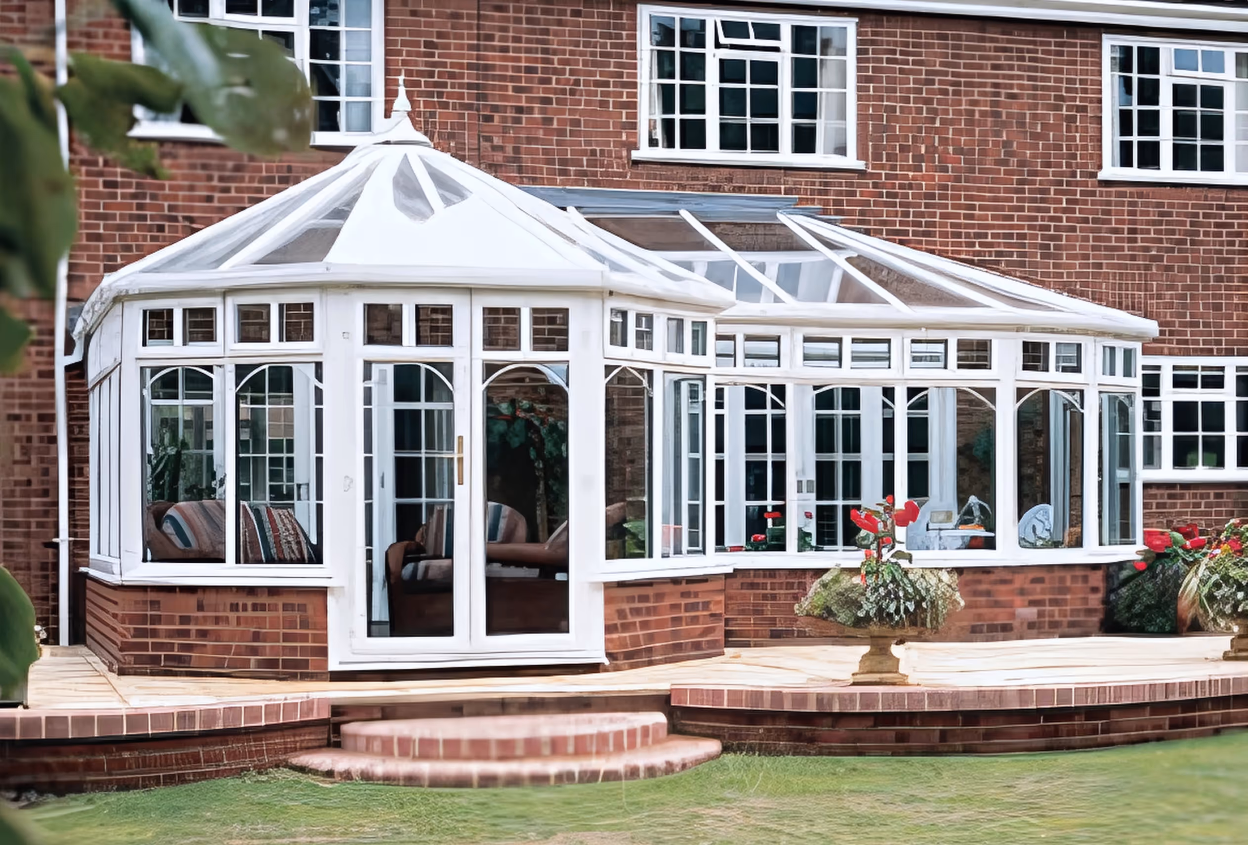 Elegant P-shaped conservatory with intricate window designs and white frames, attached to a red-brick house with garden steps leading to the entrance.
