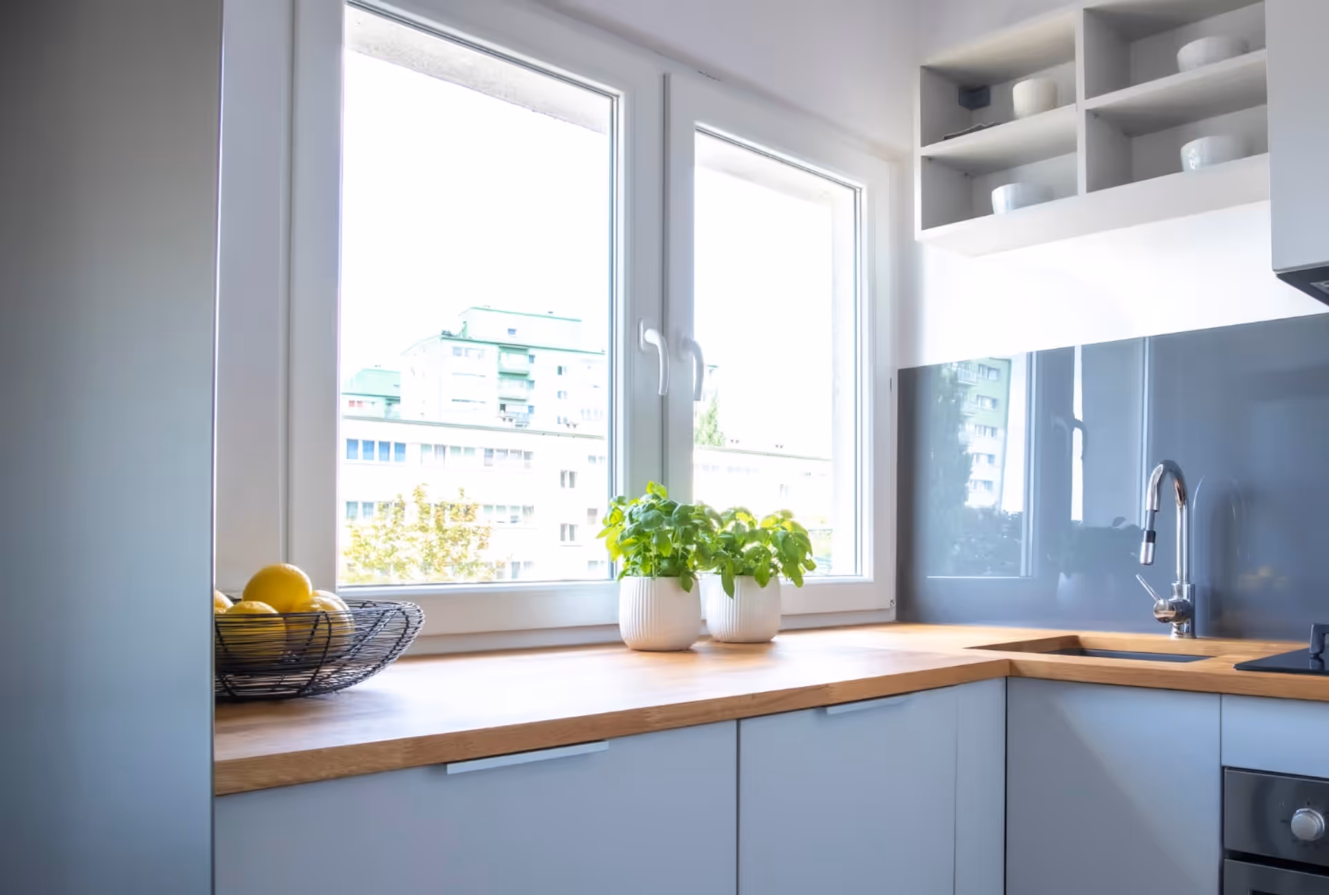 Contemporary kitchen with potted plants by the window, offering views of residential buildings under clear skies.