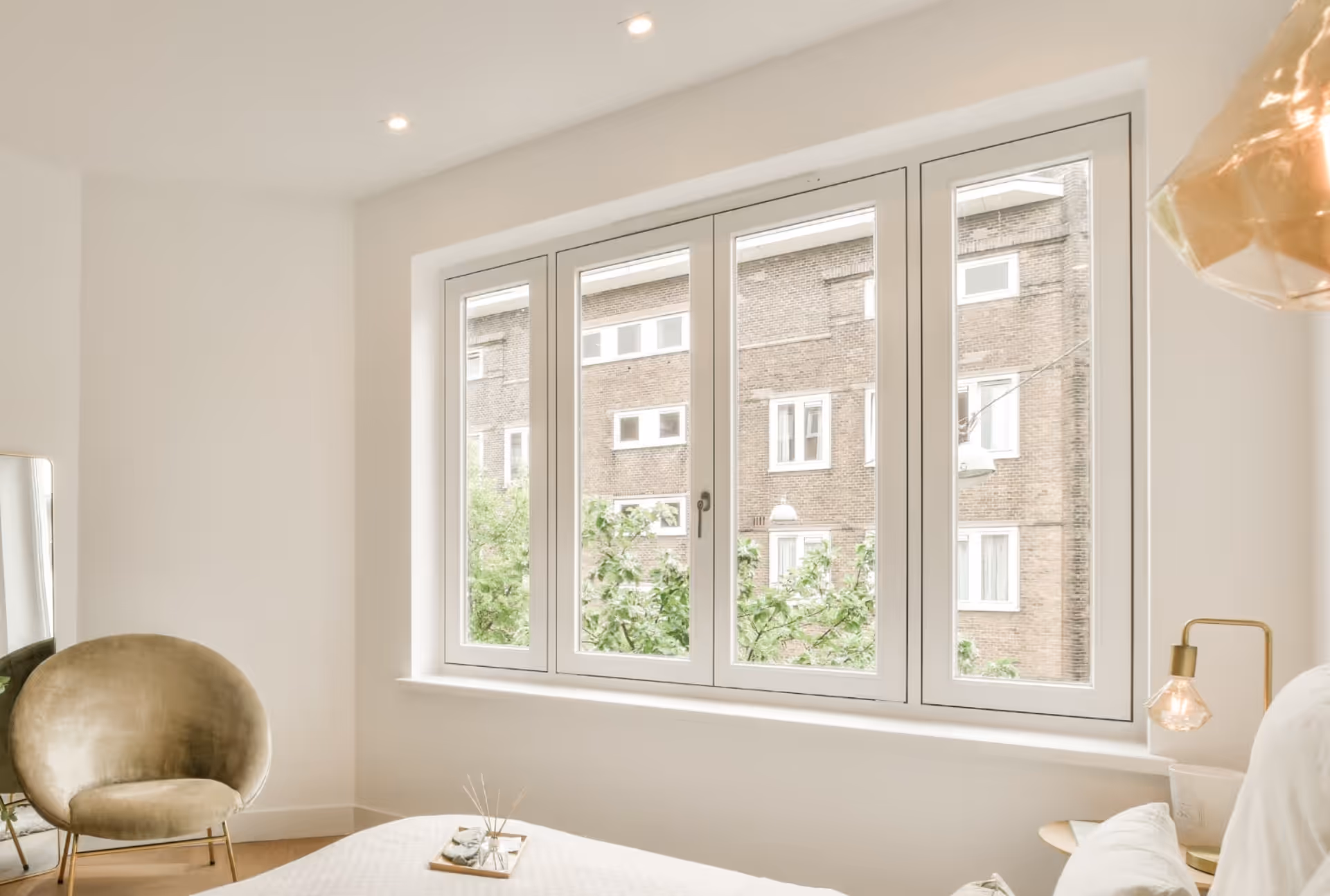 Modern bedroom with large windows showing neighbouring buildings, furnished with an armchair and bedside lamp.