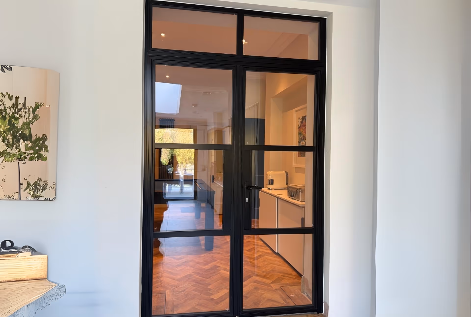 Black heritage-style glass door leading to kitchen with herringbone flooring and white walls.