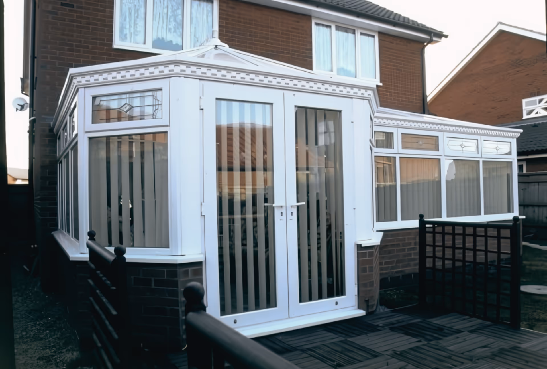 White-framed P-shaped conservatory with large glass windows and vertical blinds, attached to a brick house with decorative roofline detailing.